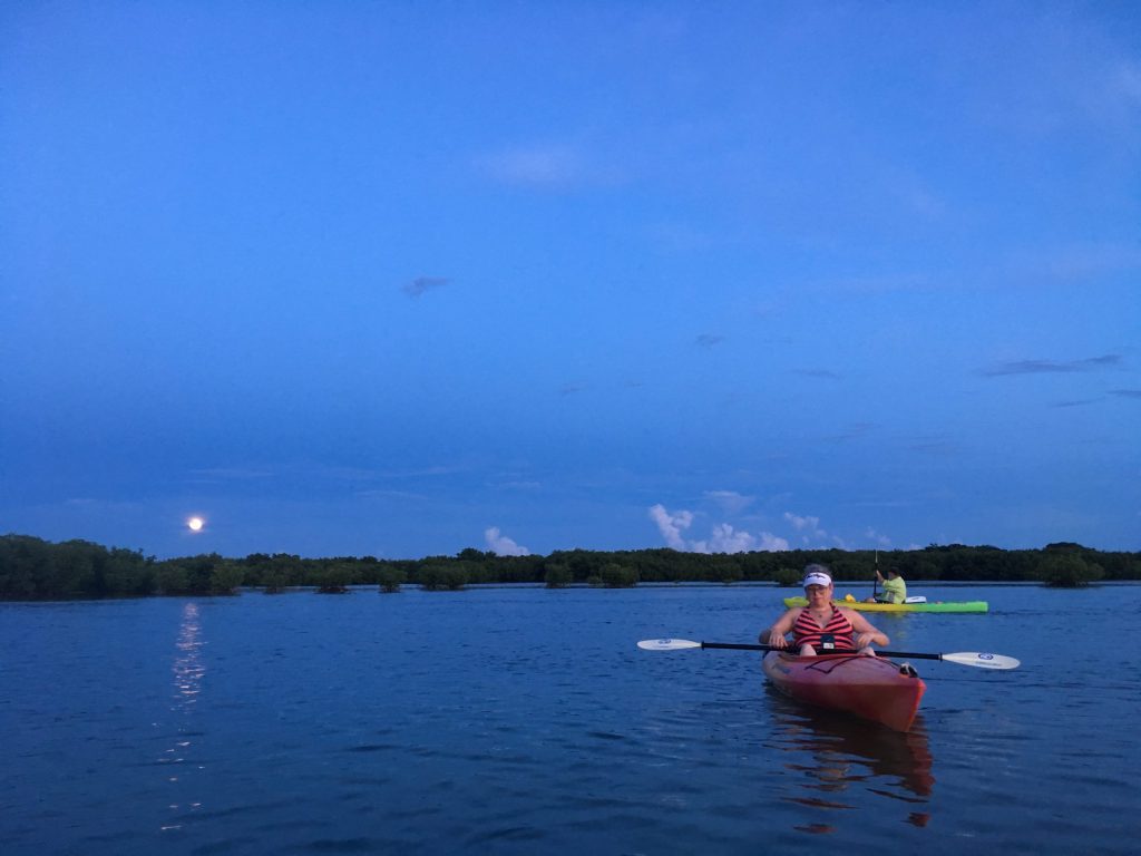 People kayaking on calm water at sunset with moon visible